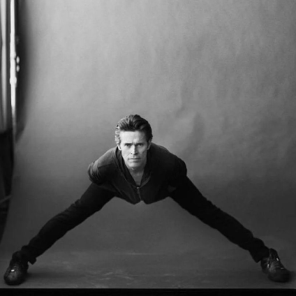 a photo of Willem Dafoe with his legs spread really wide and his torso bent forward so it looks like he is just a head on a pair of legs."