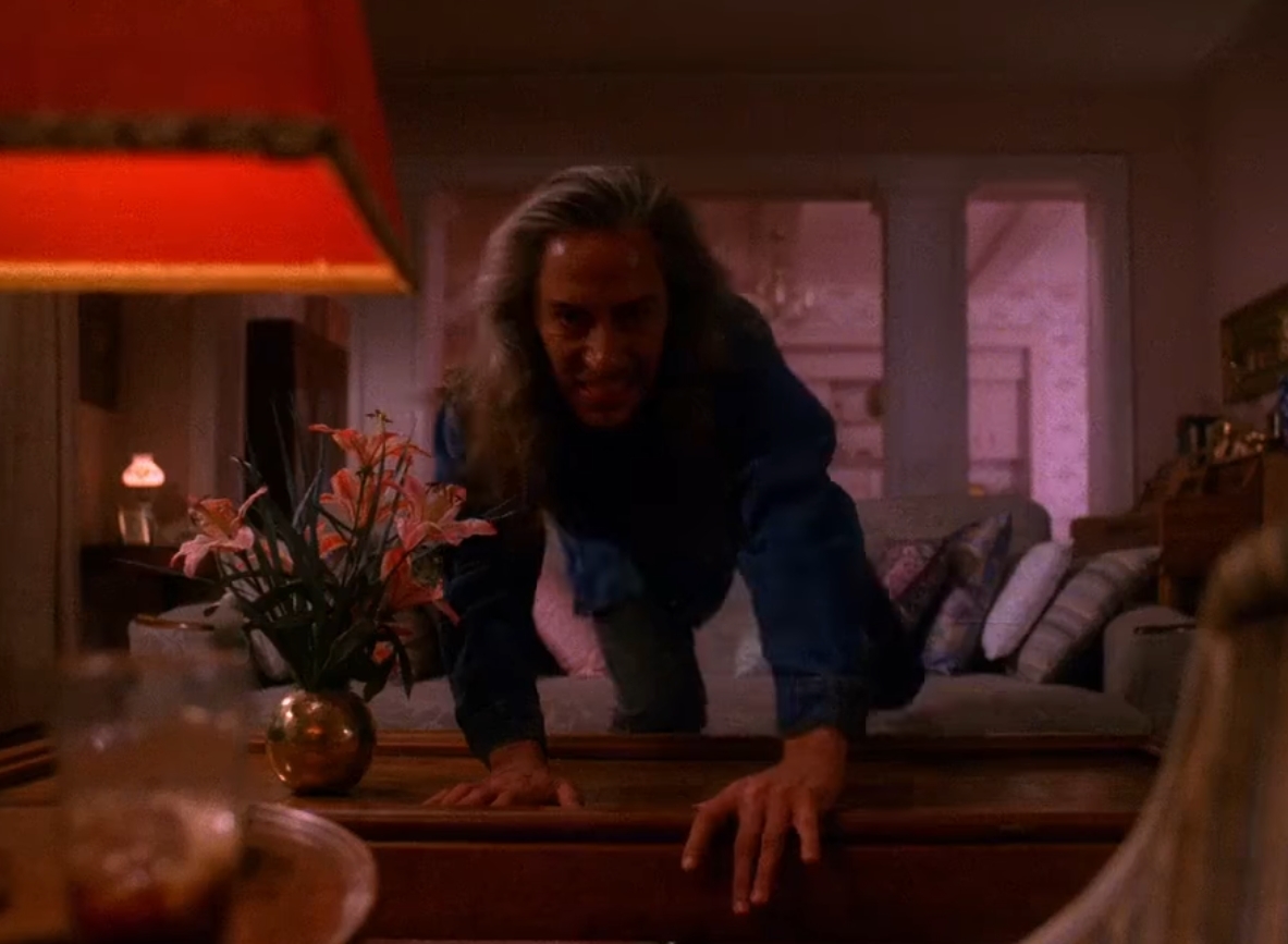 A picture of BOB from Twin Peaks. He as long grey hair and he's smiling evilly. He's climbing over a coffee table. He's looking at you and approaching you.